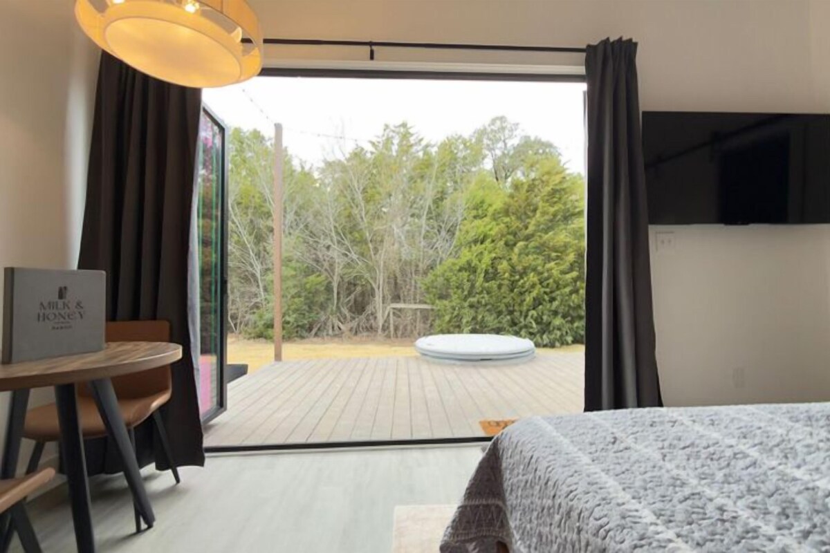 A hotel room with a bed, a table with a sign, and a view of the outdoors.