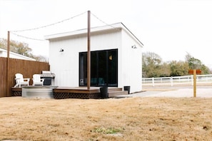 A small, single-story building with a covered patio area, a grill, and a white fence in the background.