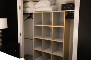 Un placard avec plusieurs étagères et une tringle pour suspendre les vêtements, un lit avec une literie blanche et une petite table de chevet avec une lampe.