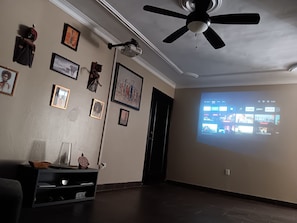 A living room with a ceiling fan, a projector screen displaying a streaming service interface, and various framed pictures on the wall. - Gazcue Executive Apartment by Peace Corps, UN, Clinics, and Universities (Santo Domingo)
