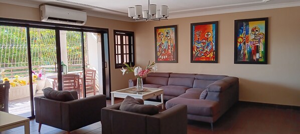 Ein Wohnzimmer mit einer Couch, zwei Sesseln, einem Couchtisch und einer Blumenvase. An der Wand hängen drei Gemälde.
