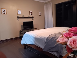 Quarto com cama, televisão sobre um suporte, janela com cortinas e quadros pendurados na parede.