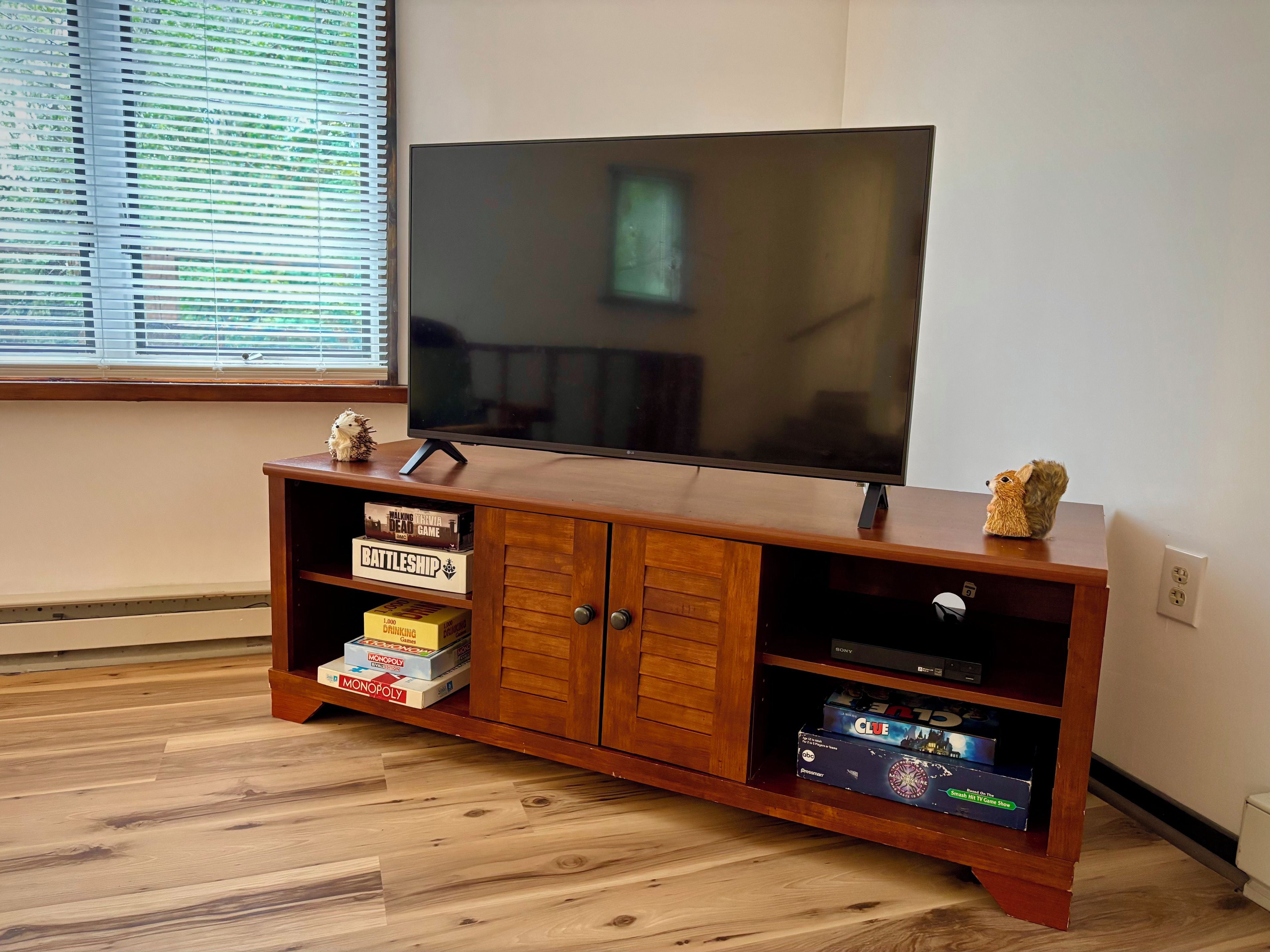 A wooden entertainment center with a flat-screen TV, shelves with board games, and a window with blinds.
