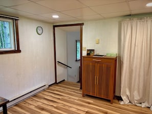 A room with a wooden floor, a wooden desk with a drawer, a clock on the wall, and a window with a view of trees.