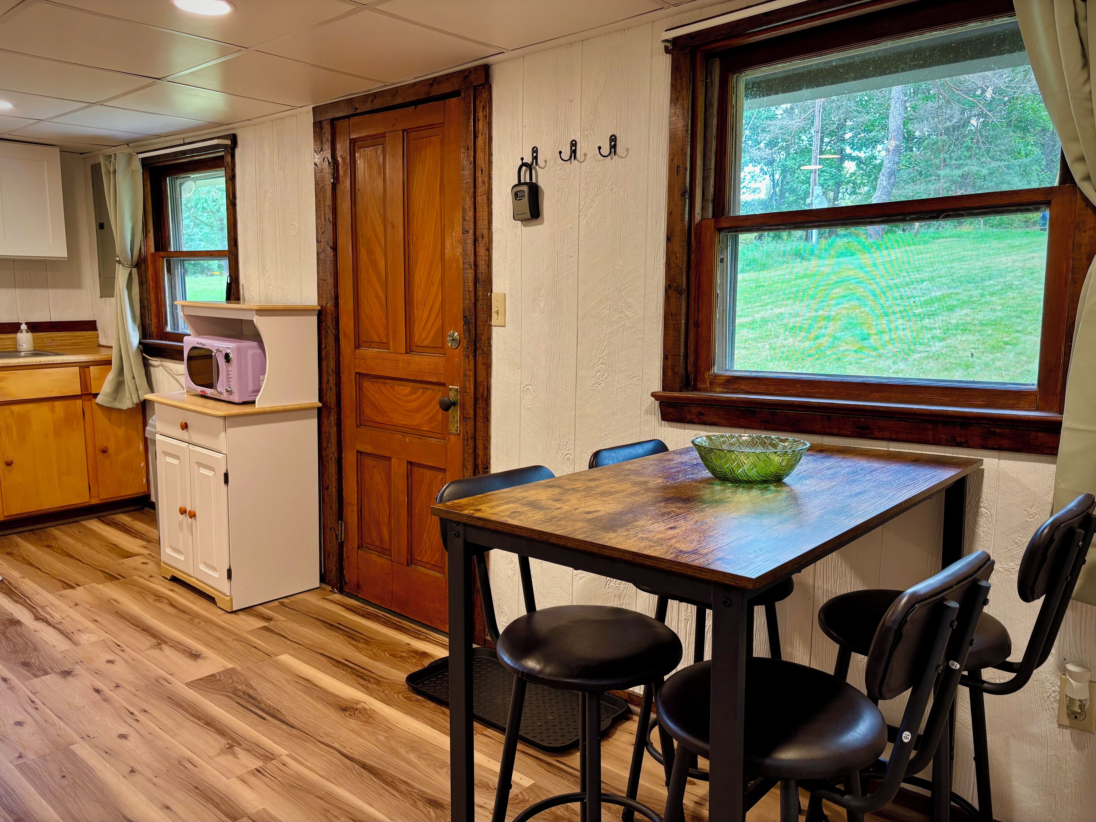 A kitchen with wooden cabinets, a microwave, a wooden door, and a window with a view of greenery.