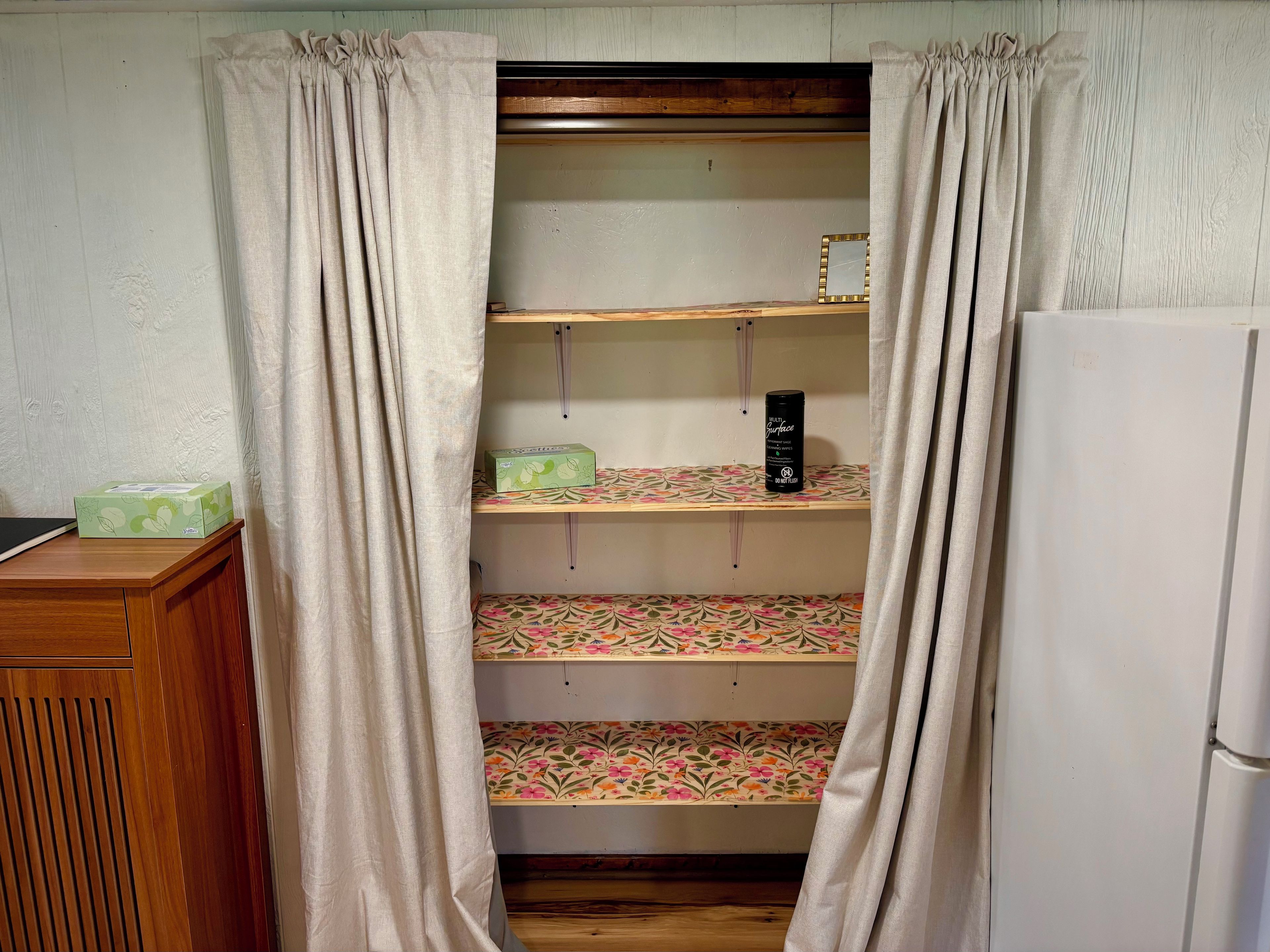 A room with a wooden cabinet, a white refrigerator, and a shelf unit with patterned fabric.