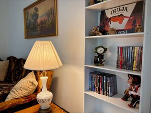 A room with a bookshelf, a lamp, a painting, and a decorative tree with a clock.