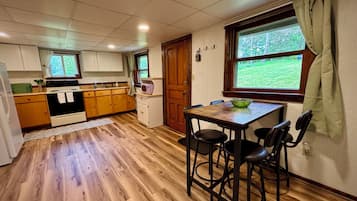 Une cuisine avec des meubles de rangement en bois, une table à manger avec des chaises, une fenêtre donnant sur un paysage verdoyant et une porte avec un cadre en bois.