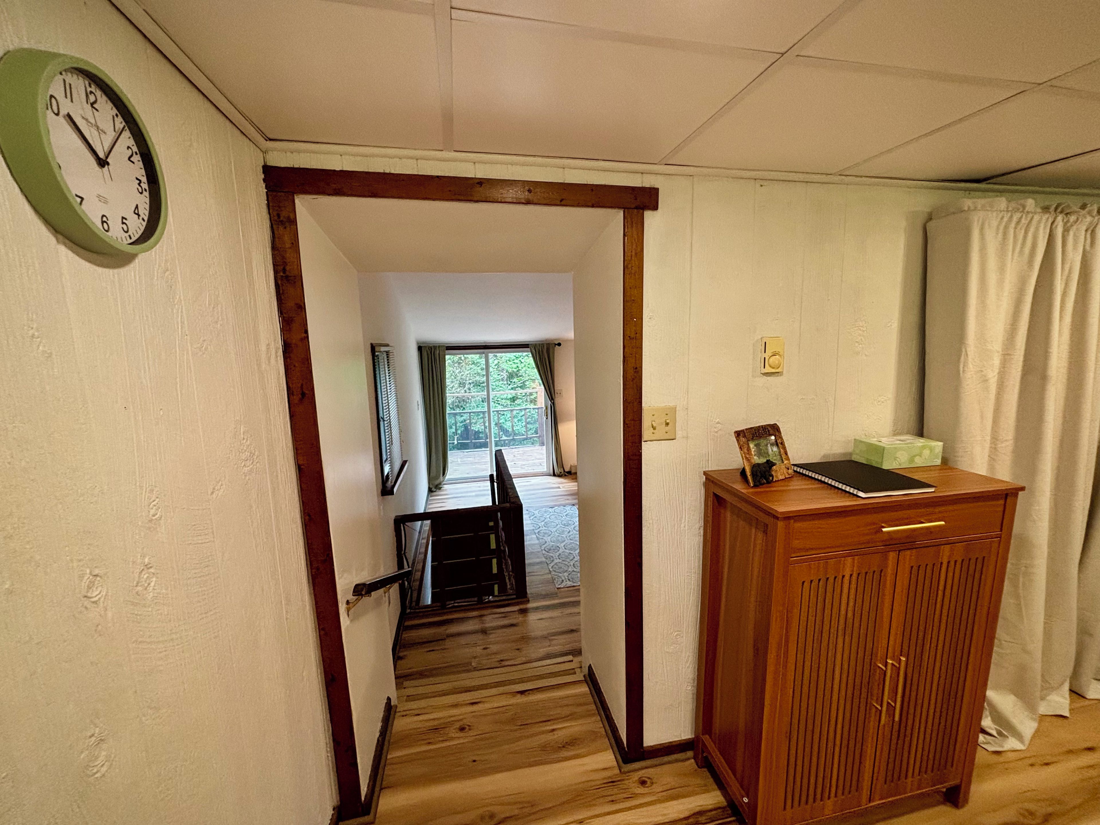 A hallway with a wooden floor, a wooden desk with a picture frame, a clock, and a window with curtains.