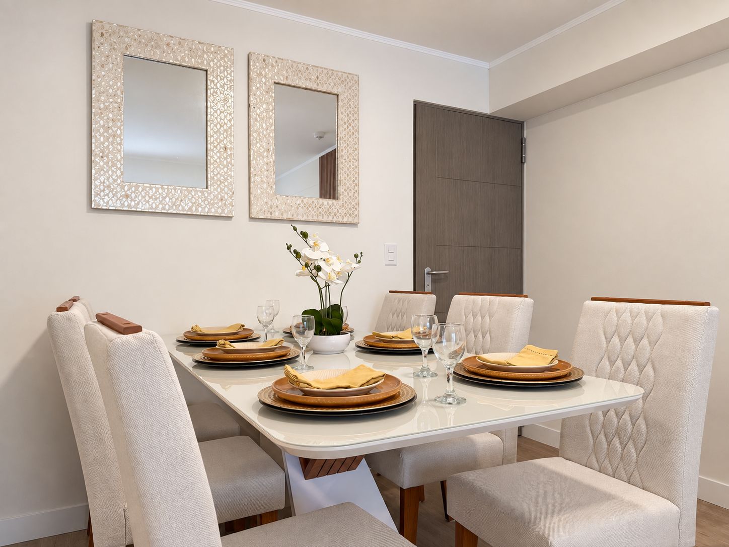 A dining room with a rectangular table set for four, white upholstered chairs, two wall-mounted mirrors, and a door in the background.