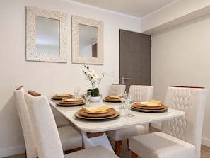 A dining room with a rectangular table set for four, white upholstered chairs, two wall-mounted mirrors, and a door in the background. - Modern apartment with terrace in Miraflores (101) (Miraflores)
