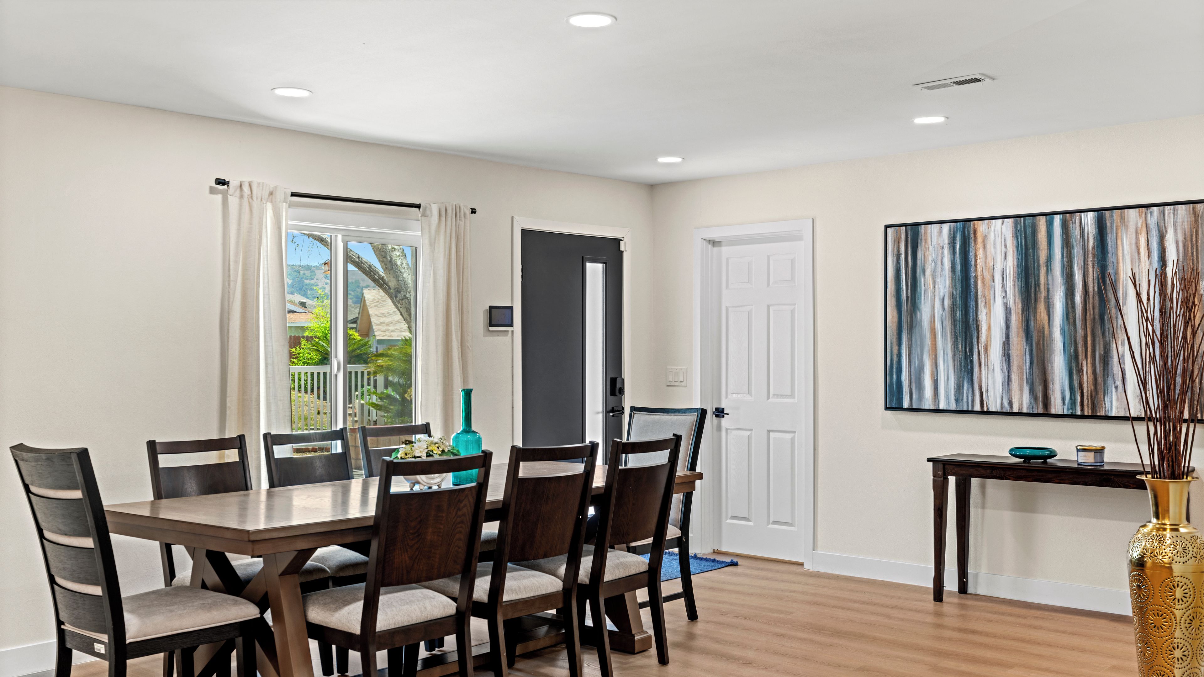 A dining area with a wooden table and chairs, a door, a window with curtains, and a painting on the wall.