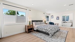 Une chambre à coucher moderne avec un grand lit, des tables de chevet, un fauteuil, une coiffeuse avec une baignoire et des œuvres d’art accrochées aux murs.