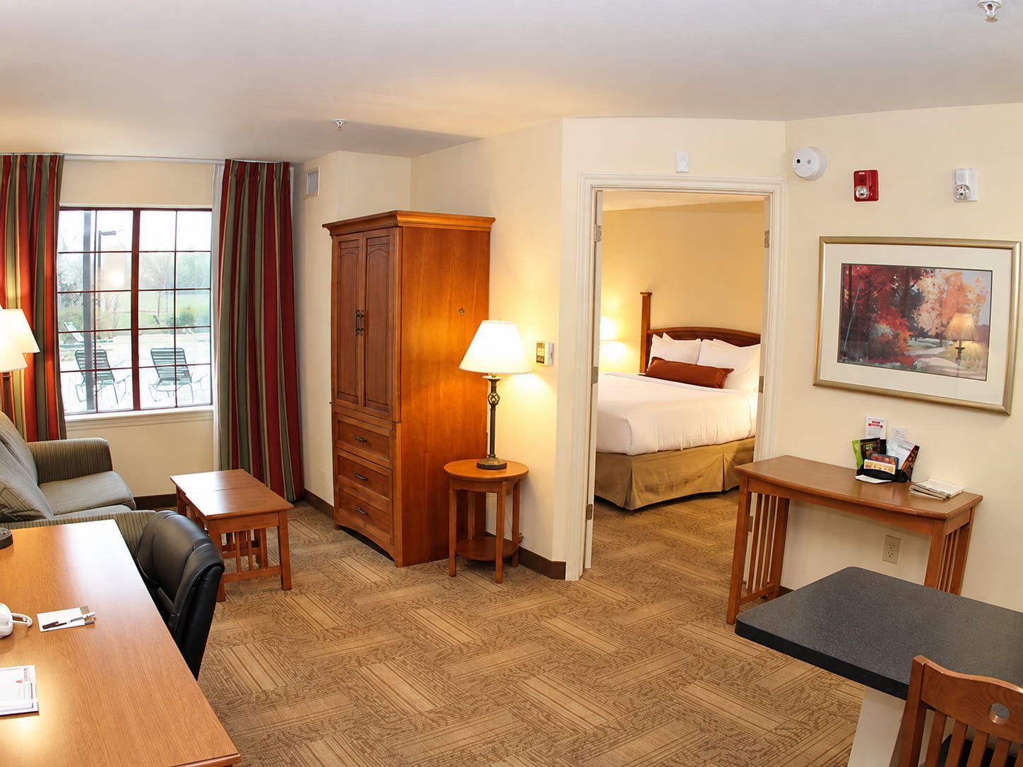 A hotel room with a bed, a desk, a chair, a wardrobe, a lamp, a painting, and a window with curtains.