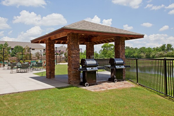A covered outdoor area with stone pillars, two barbecue grills, and a fenced patio with chairs and tables.