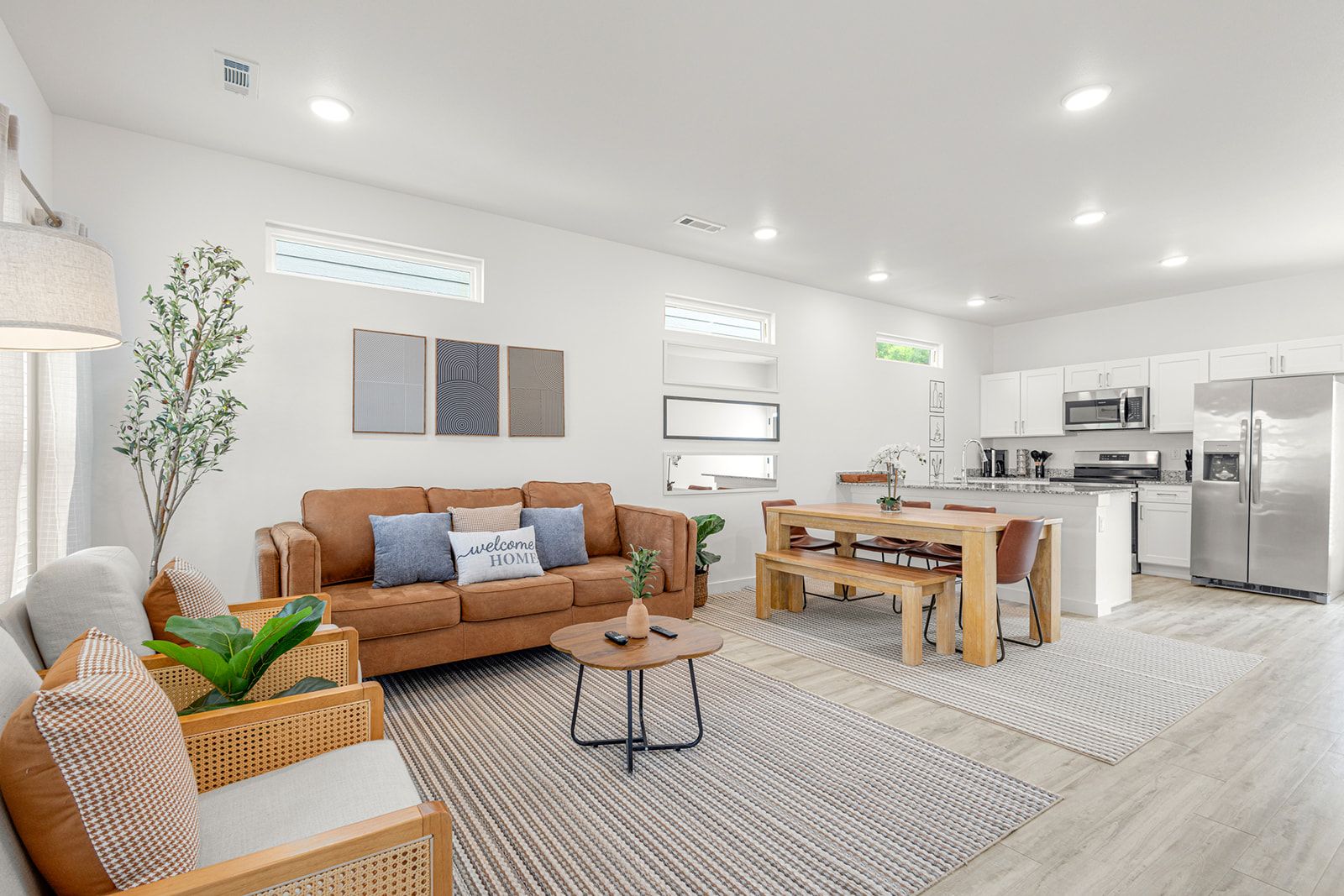 A modern living room with a sofa, coffee table, and dining table. There is a kitchen area with appliances and a refrigerator.