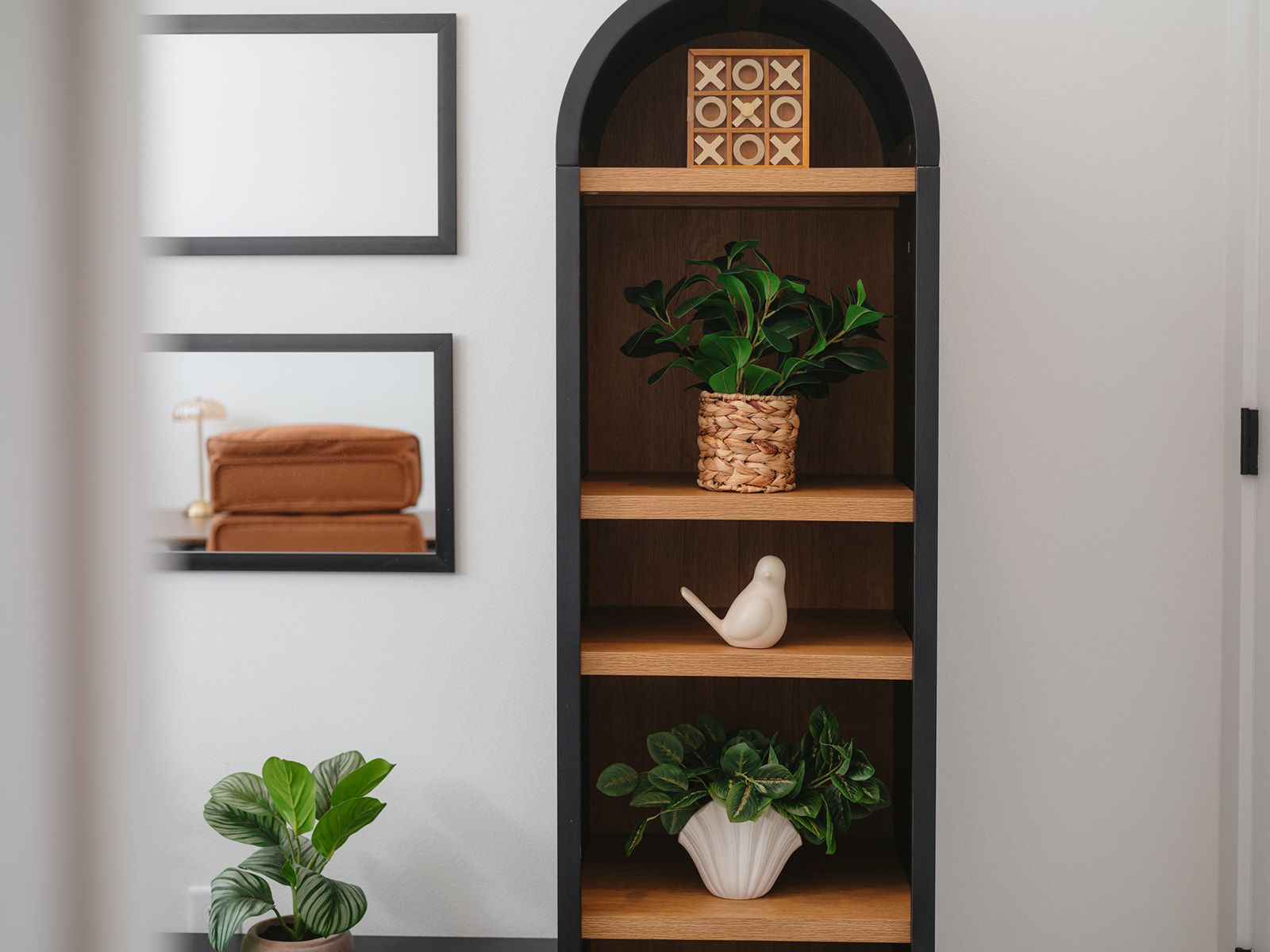 Una pared con un espejo de marco negro, un estante de madera con una cesta tejida y una escultura de pájaro blanco.