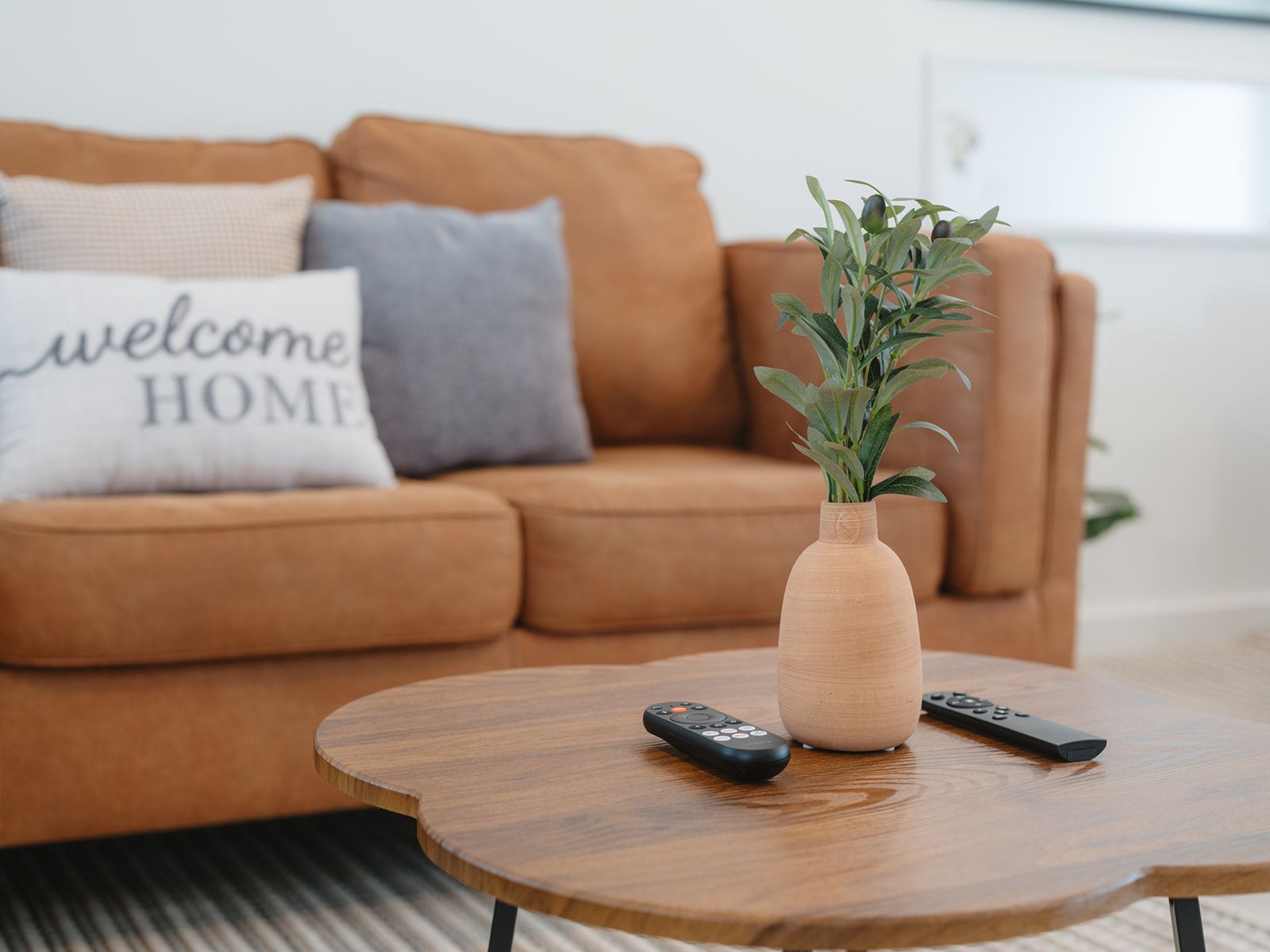 Una sala de estar con un sofá, una mesa redonda de madera, un jarr ón con plantas verdes y dos controles remotos.