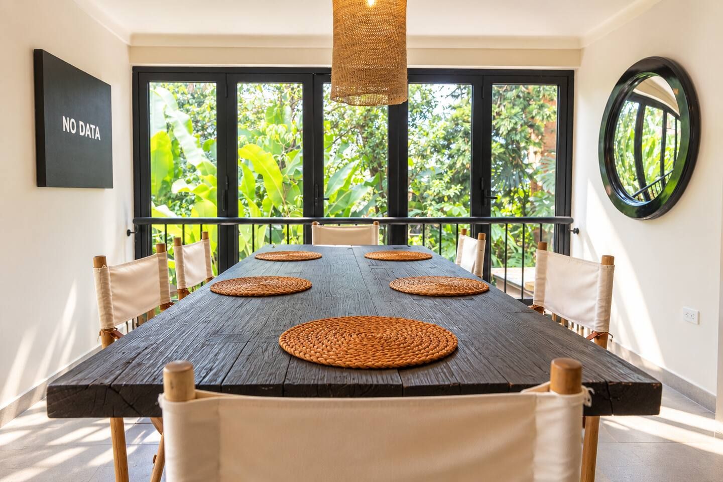 Un comedor con una gran mesa de madera, individuales tejidos y sillas blancas con cojines.