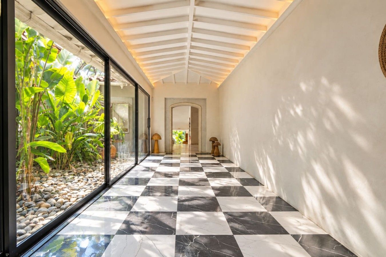 Un pasillo con piso de cuadros, una pared de vidrio con plantas y un plafón con lámpara.