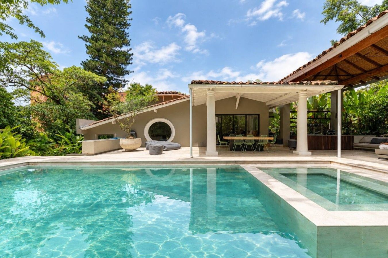 Una piscina cristalina con un porche techado, mobiliario exterior y abundante vegetación.
