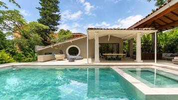 Una piscina cristalina con un porche techado, mobiliario exterior y abundante vegetación.