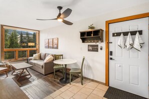 Een moderne woonkamer met een bank, een ronde tafel en stoelen. Er is een plafondventilator, een raam met uitzicht op bomen, en een deur met handdoeken aan haken.