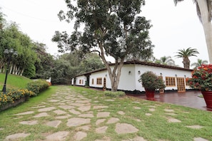 A white building with a tiled roof, surrounded by a garden with a stone pathway. - Cozy 12BR House with Pool in Pachacamac (Pachacámac)