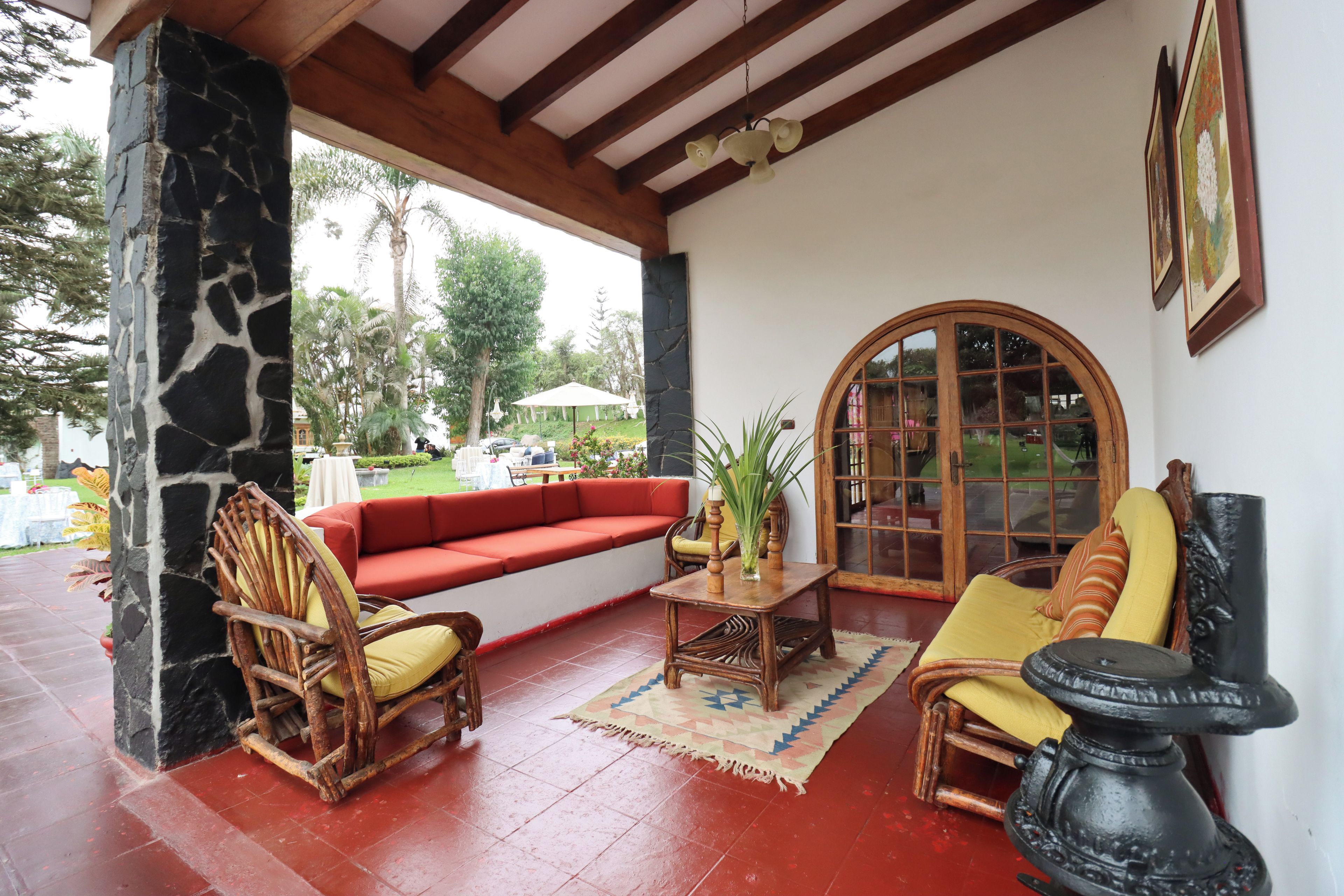 A covered outdoor seating area with a stone pillar, a red sofa, wooden chairs, a table with a vase, and a large window.
