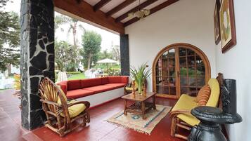 A covered outdoor seating area with a stone pillar, a red sofa, wooden chairs, a table with a vase, and a large window.