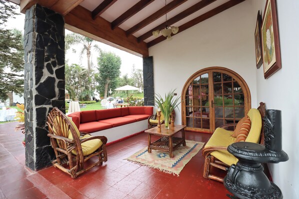 A covered outdoor seating area with a stone pillar, a red sofa, wooden chairs, a table with a vase, and a large window.