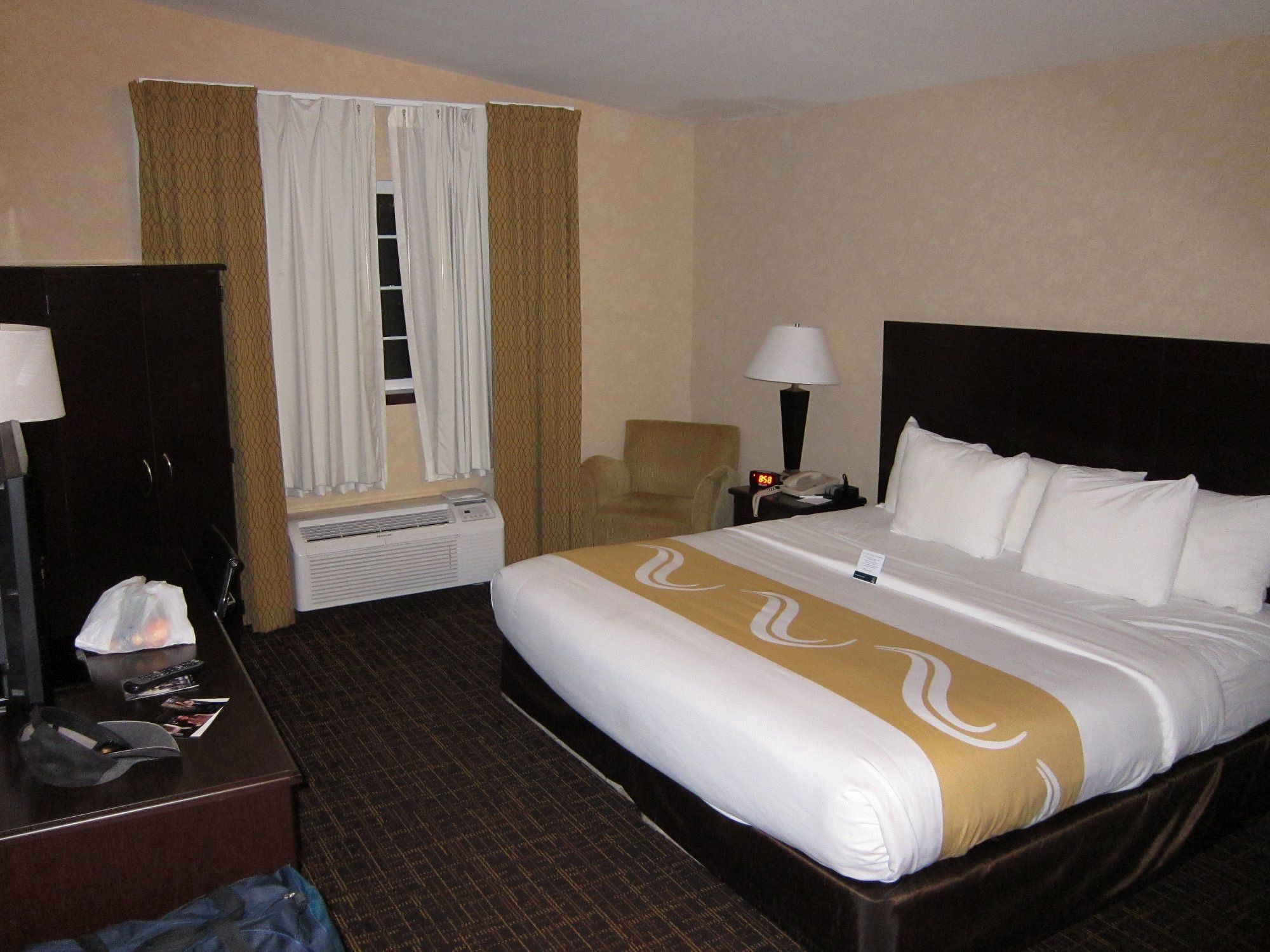 Quarto de hotel com cama, escrivaninha, cadeira, mesa de cabeceira com luminária e janela com cortinas.