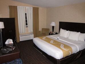 Quarto de hotel com cama, escrivaninha, cadeira, mesa de cabeceira com luminária e janela com cortinas.