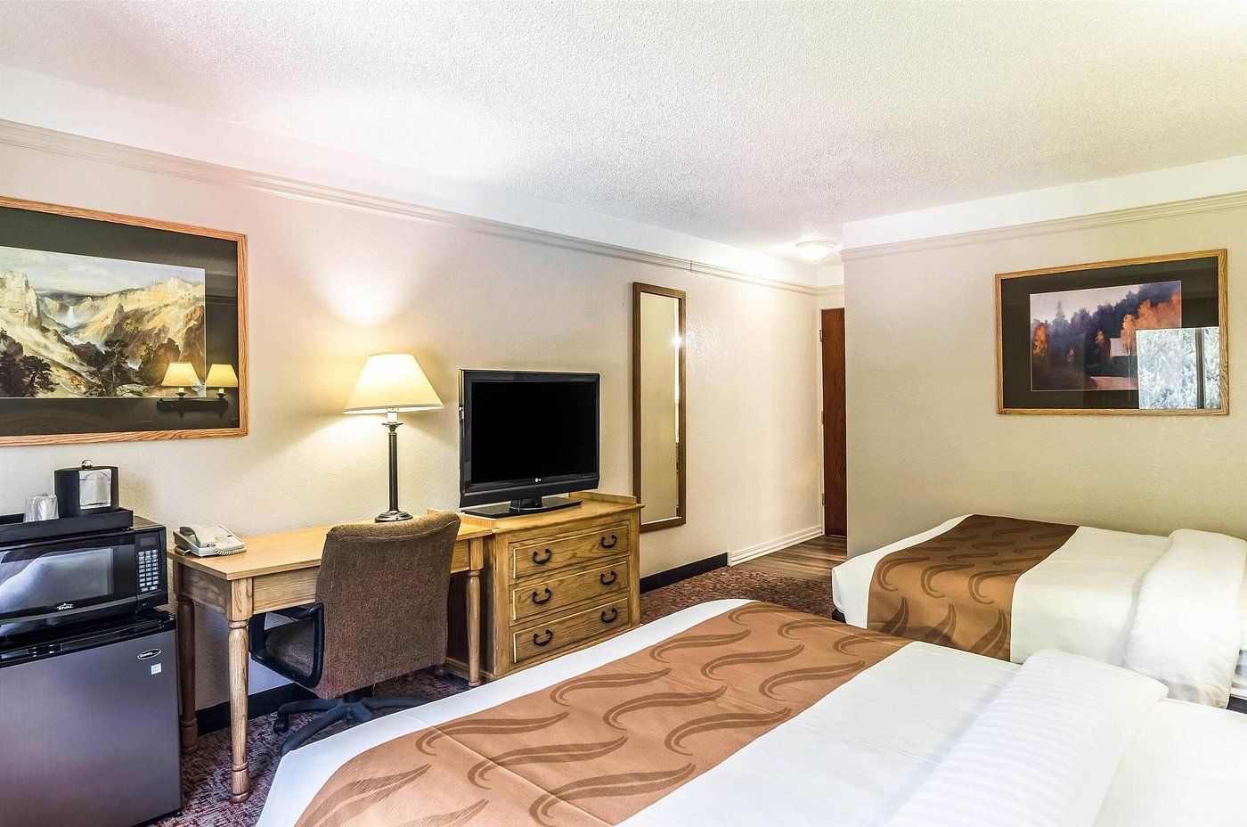 A hotel room with a desk, television, two beds, a chair, and a refrigerator.