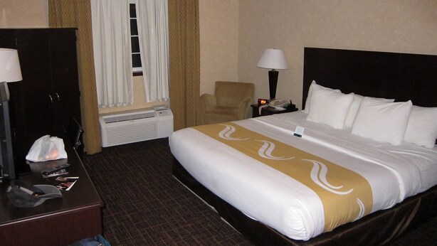 Una camera d'albergo con un letto, una scrivania, una sedia, un comodino con una lampada e una finestra con tende.
