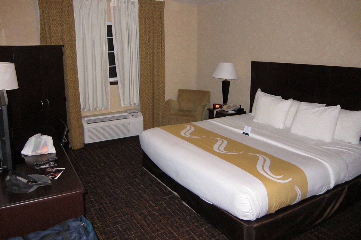 Quarto de hotel com cama, escrivaninha, cadeira, mesa de cabeceira com luminária e janela com cortinas.