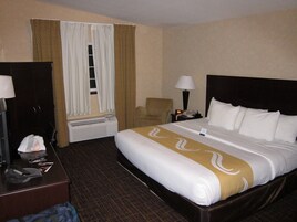 Habitación de hotel con cama, escritorio, silla, mesita de noche con lámpara y ventana con cortinas.