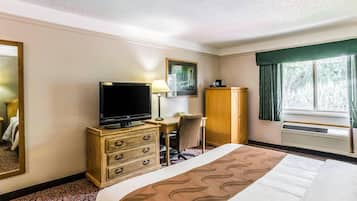 Quarto de hotel com cama, escrivaninha com cadeira, televisão, guarda-roupa e janela com cortinas.
