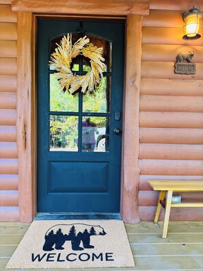 A wooden cabin door with a wreath, a welcome sign, and a doormat with a bear silhouette. - 3 mile scenic ride to WPG trails from Sunset Vista (Mena)