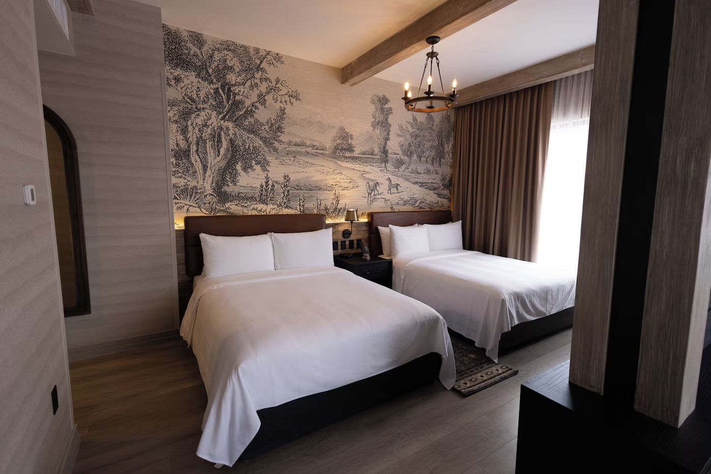 Una habitación de hotel con dos camas, un mural grande en la pared, una araña y una ventana con cortinas.