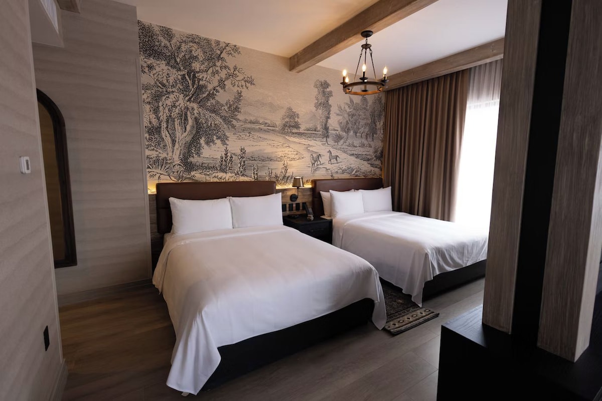 A hotel room with two beds, a large mural on the wall, a chandelier, and a window with curtains.