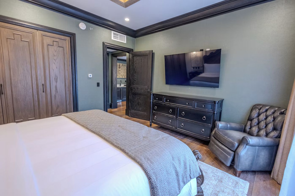 A bedroom with a large bed, a dark wooden dresser, a leather armchair, and a flat-screen TV on the wall.