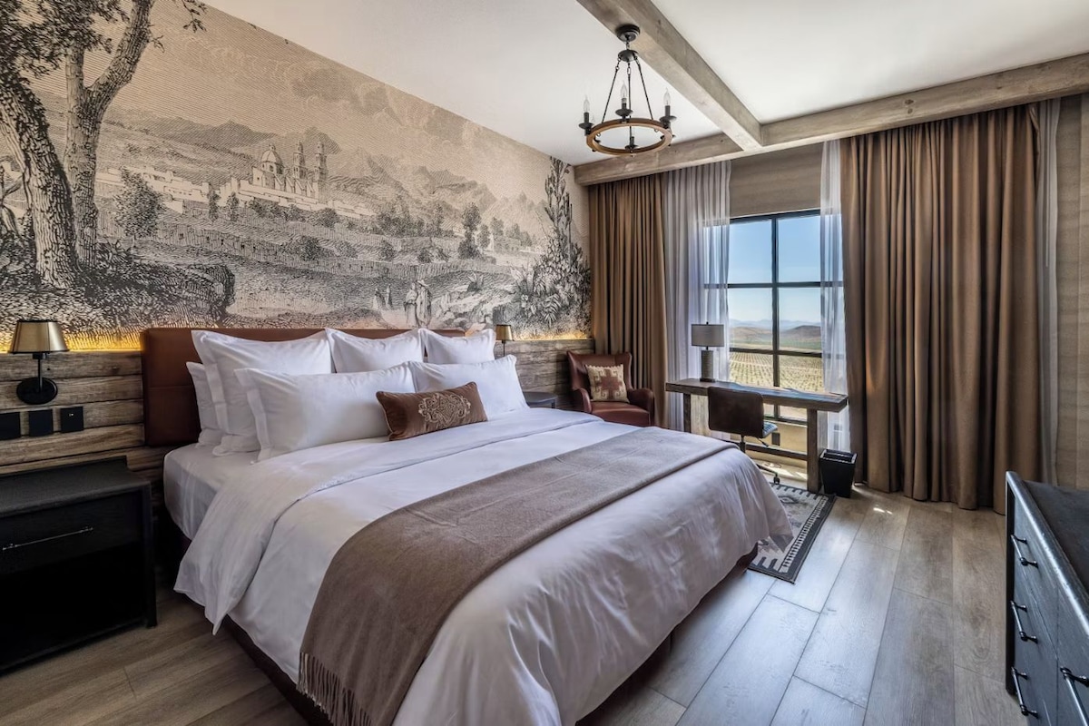 A hotel room with a large bed, a view of the desert through the window, a chandelier, and a mural on the wall.