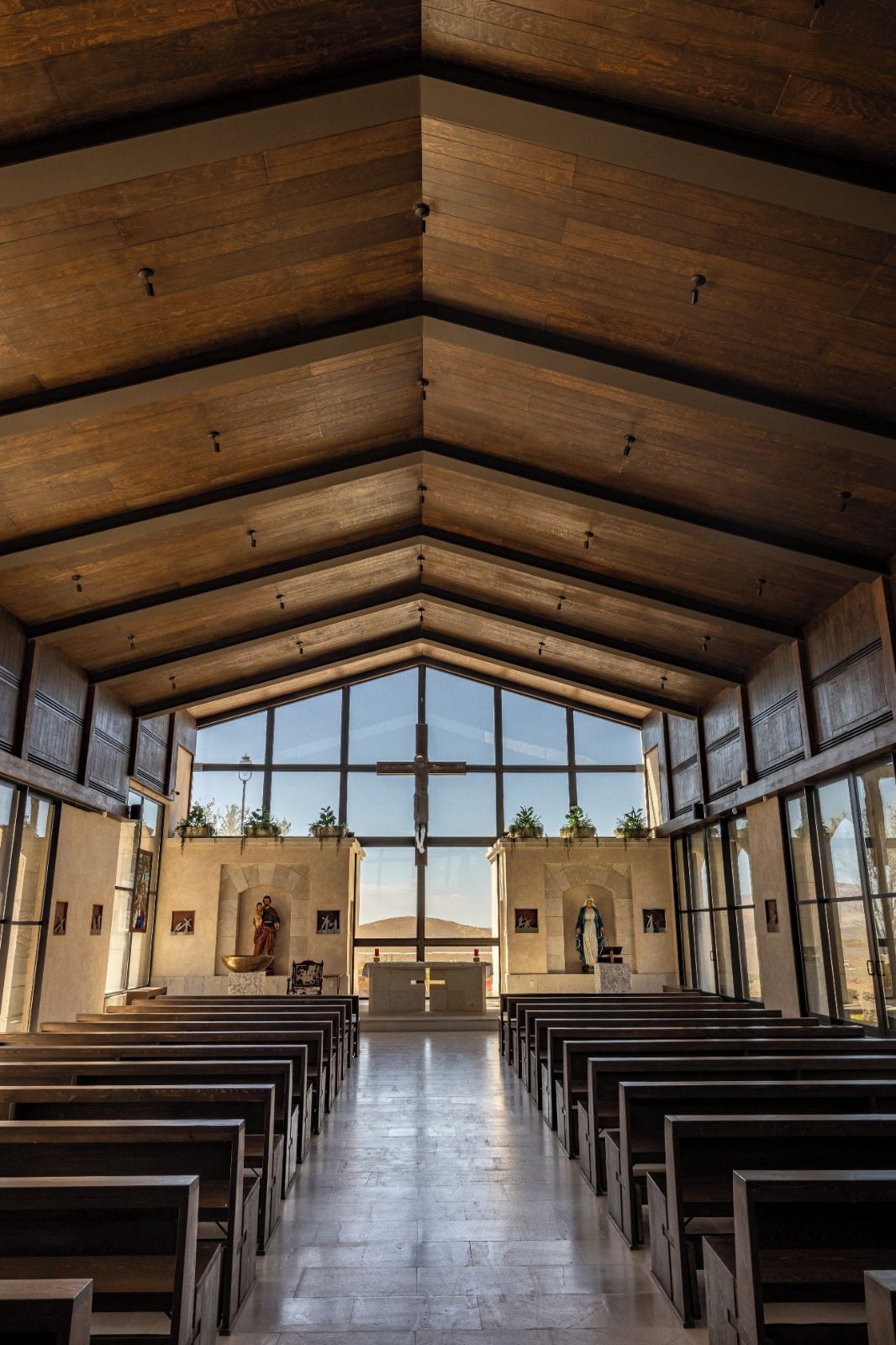 L’intérieur d’une église avec des bancs en bois, un crucifix et une grande fenêtre en arc de cercle.