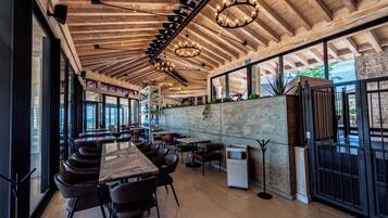 Un restaurante con una barra alargada, varias mesas y sillas. Hay lámparas colgantes y vistas al exterior a través de amplias ventanas.