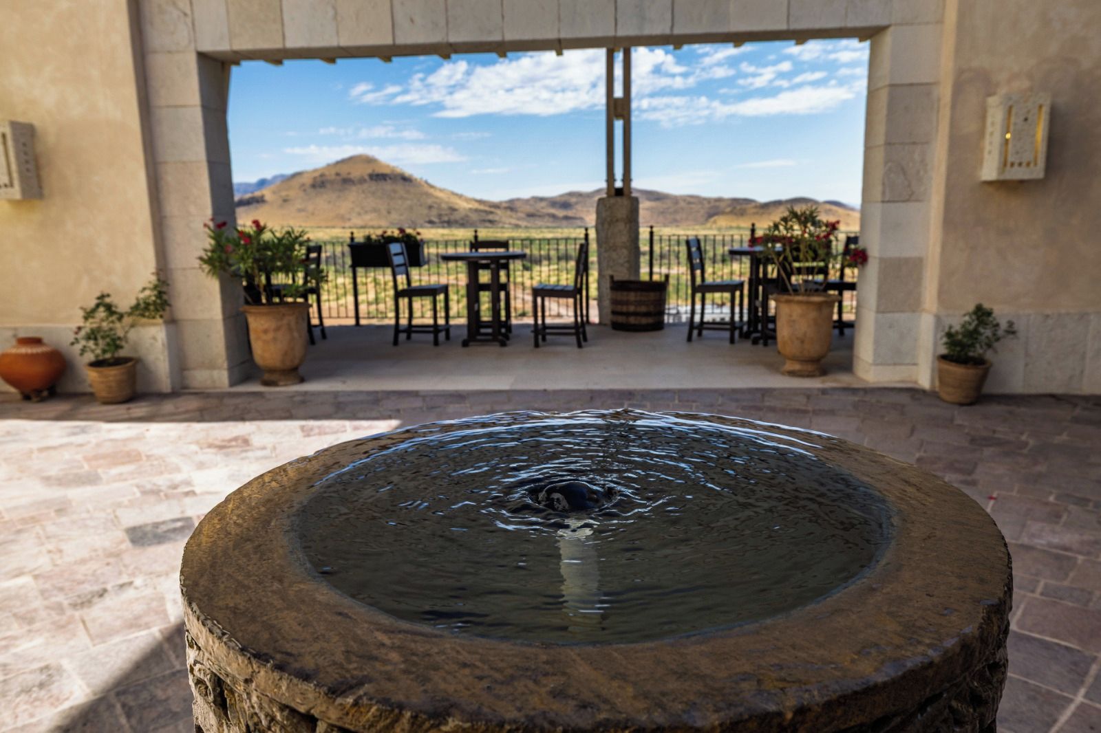 A stone fountain in the foreground with a patio area featuring tables and chairs, potted plants, and a view of a mountain range in the background.