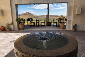 A stone fountain in the foreground with a patio area featuring tables and chairs, potted plants, and a view of a mountain range in the background.