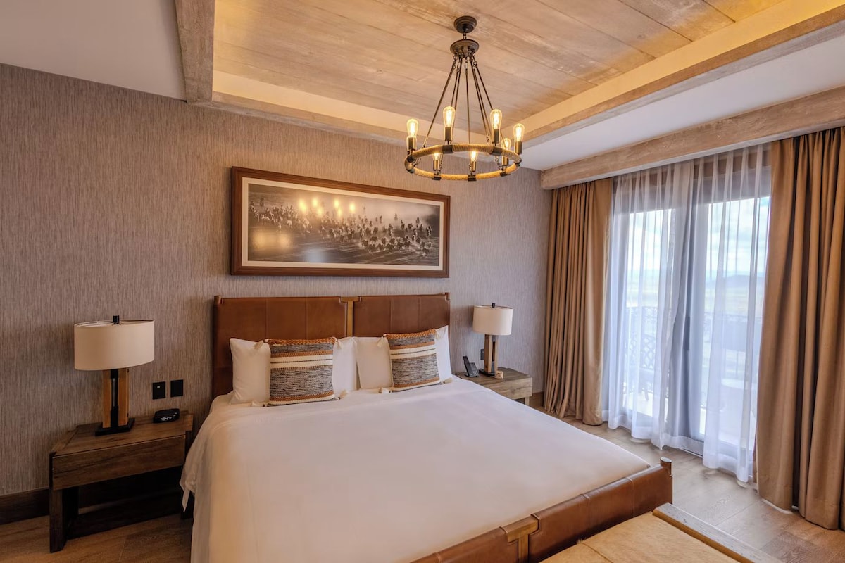 A hotel room with a large bed, a chandelier, a painting on the wall, and a view through the window.