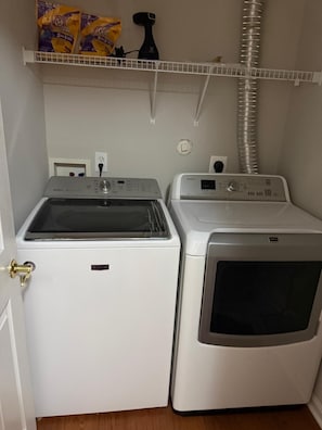 A laundry room with a front-loading washer and dryer, a shelf with cleaning supplies, and a vent hose.