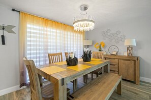 A dining room with a wooden table, chairs, a chandelier, and a window with curtains. - 3BR Screened Heated Saltwater Pool • BBQ (Port Richey)